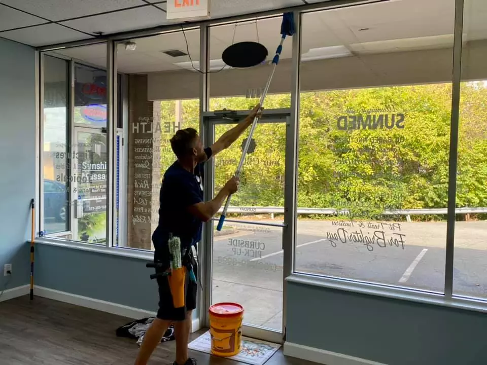 A worker is cleaning the large glass windows of a house cleaning business storefront with a long-handled tool, standing inside the shop near an orange bucket, while the street and green trees are visible outside.