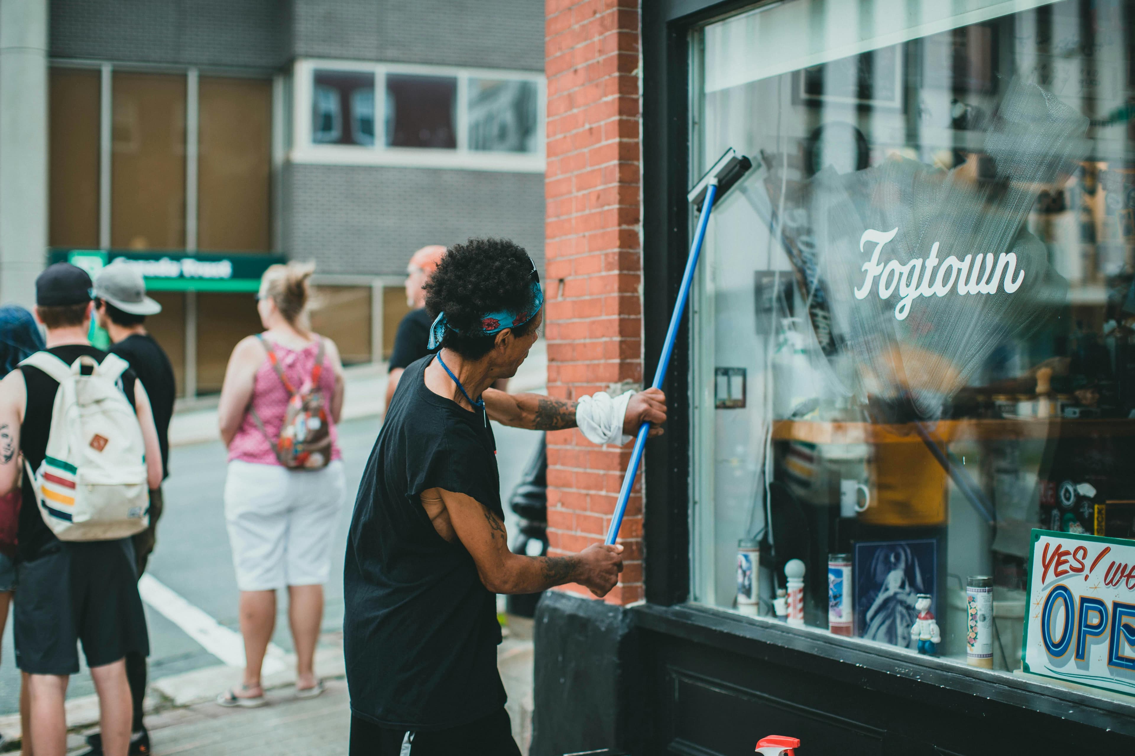 A person with curly hair and tattoos cleaning the window of a house cleaning business storefront named "Fogtown".