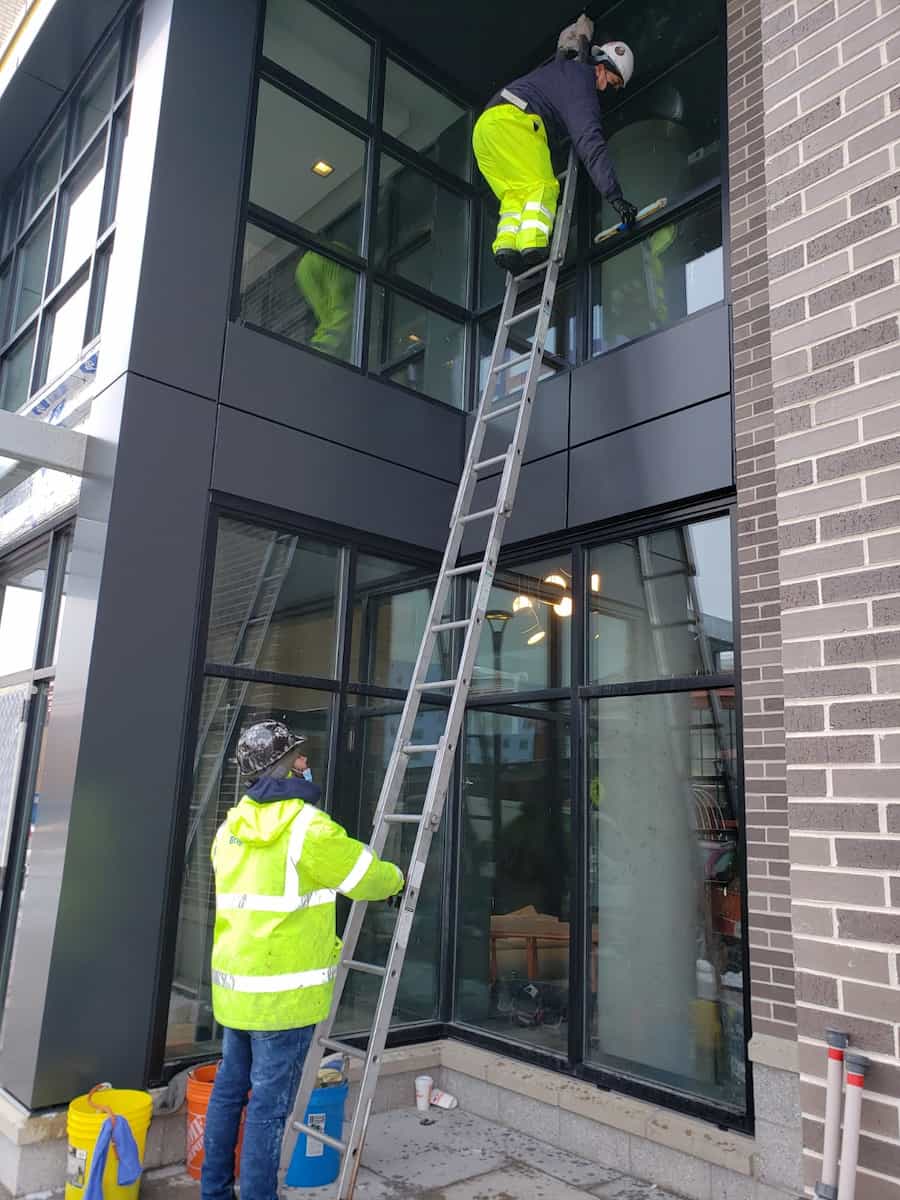 Two workers in safety gear are cleaning the exterior windows of a modern multi-story building using a ladder and cleaning tools.